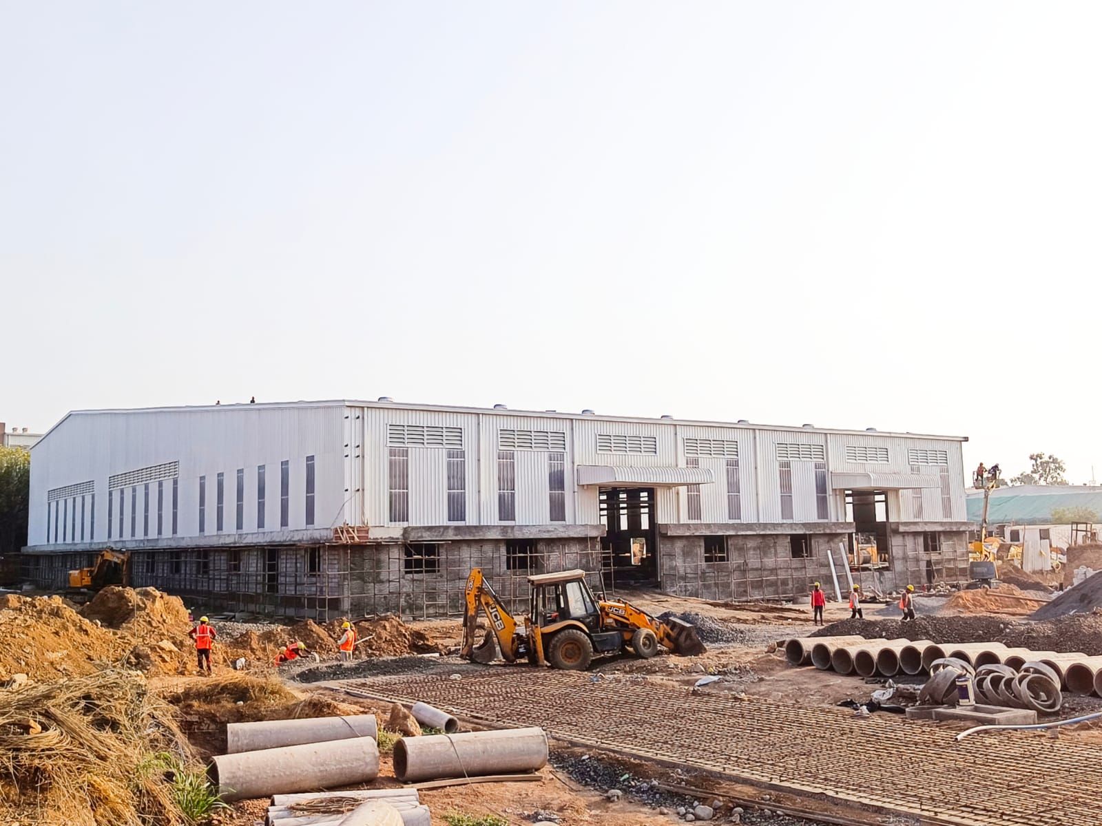 Large white PEB factory building with workers and a backhoe loader doing site works in front